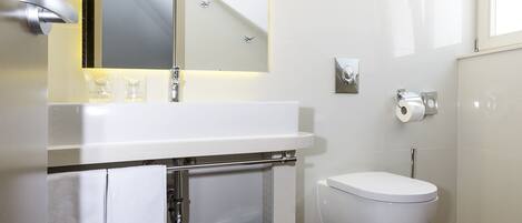 A modern bathroom with a white sink, mirror, and toilet. There is a towel rack with towels, a trash bin, and a small mat on the floor.