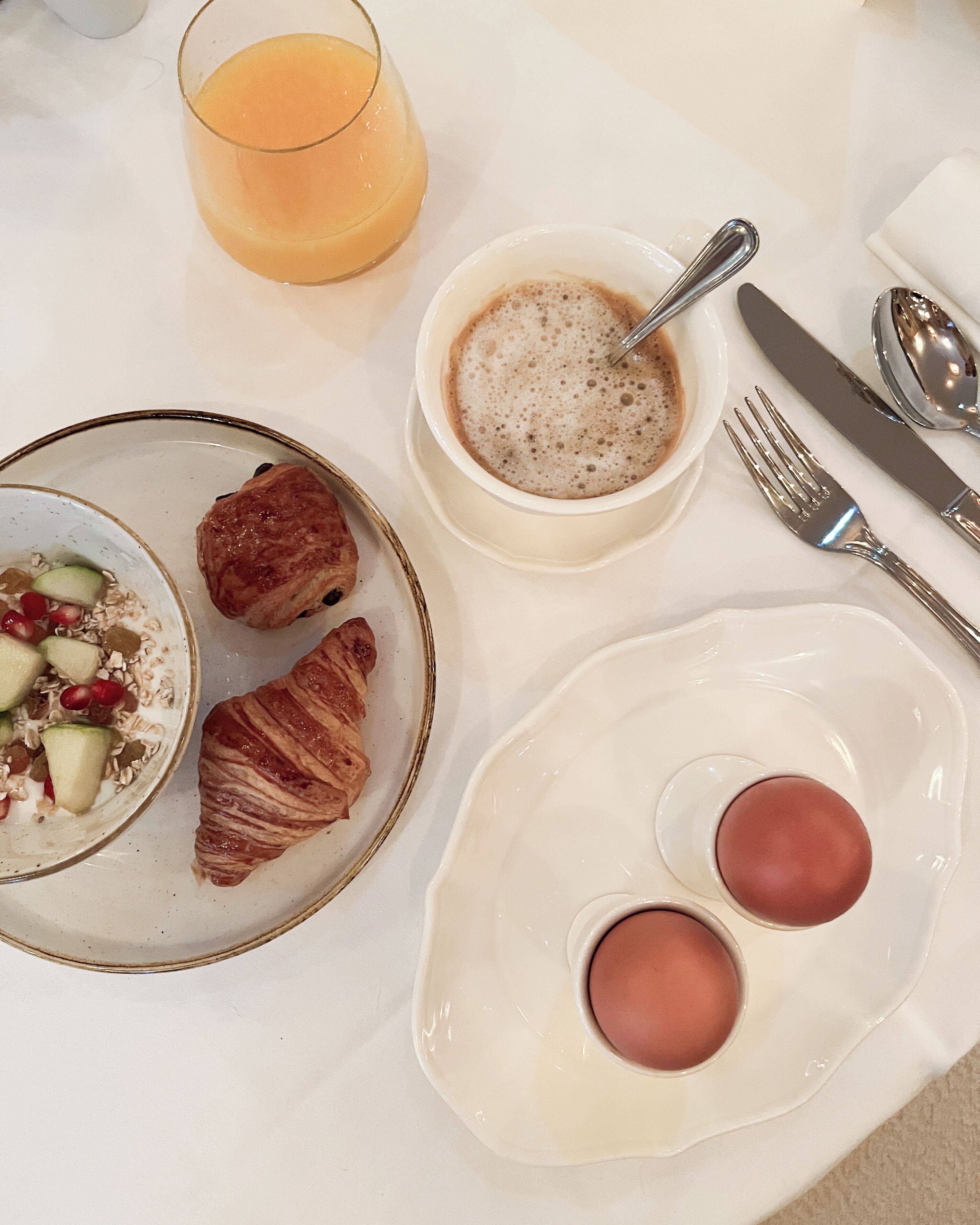 A breakfast setting with croissants, a bowl of coffee, a glass of orange juice, and a plate with eggs.