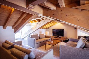 Een moderne loft-woonkamer met zichtbare houten balken, een grote flatscreen televisie en een gezellige zithoek met banken en een salontafel.