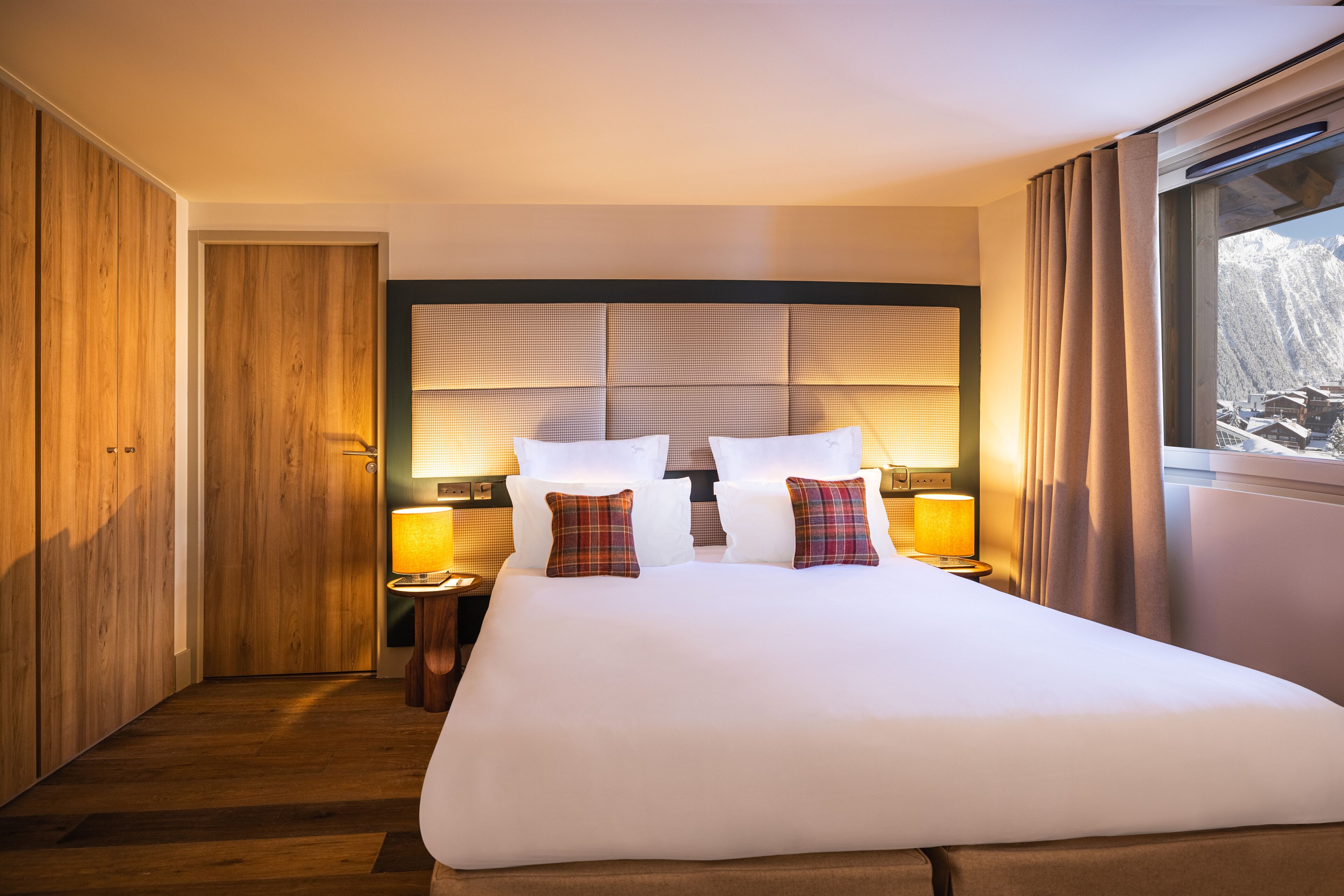 A hotel room with a large bed, wooden wardrobe, and a view of mountains through the window.
