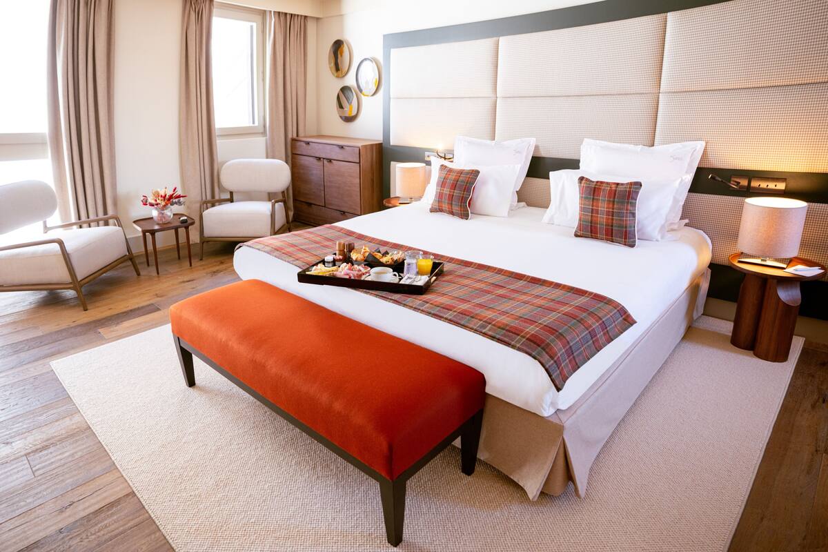 Een hotelkamer met een groot bed, een nachtkastje met een lamp, een stoel, een klein tafeltje met een vaas, en een raam met gordijnen.