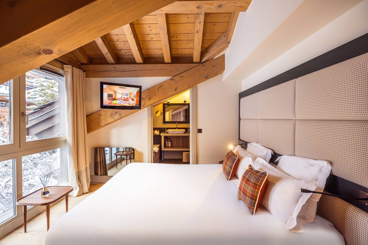 Een slaapkamer met een groot bed, houten balken in het plafond, een televisie en uitzicht op het omliggende landschap.