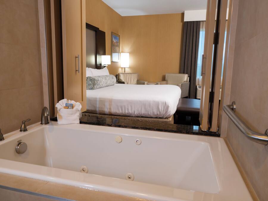 A hotel room with a large bathtub, a bed with white linens, a bedside table with lamps, and a mirror.