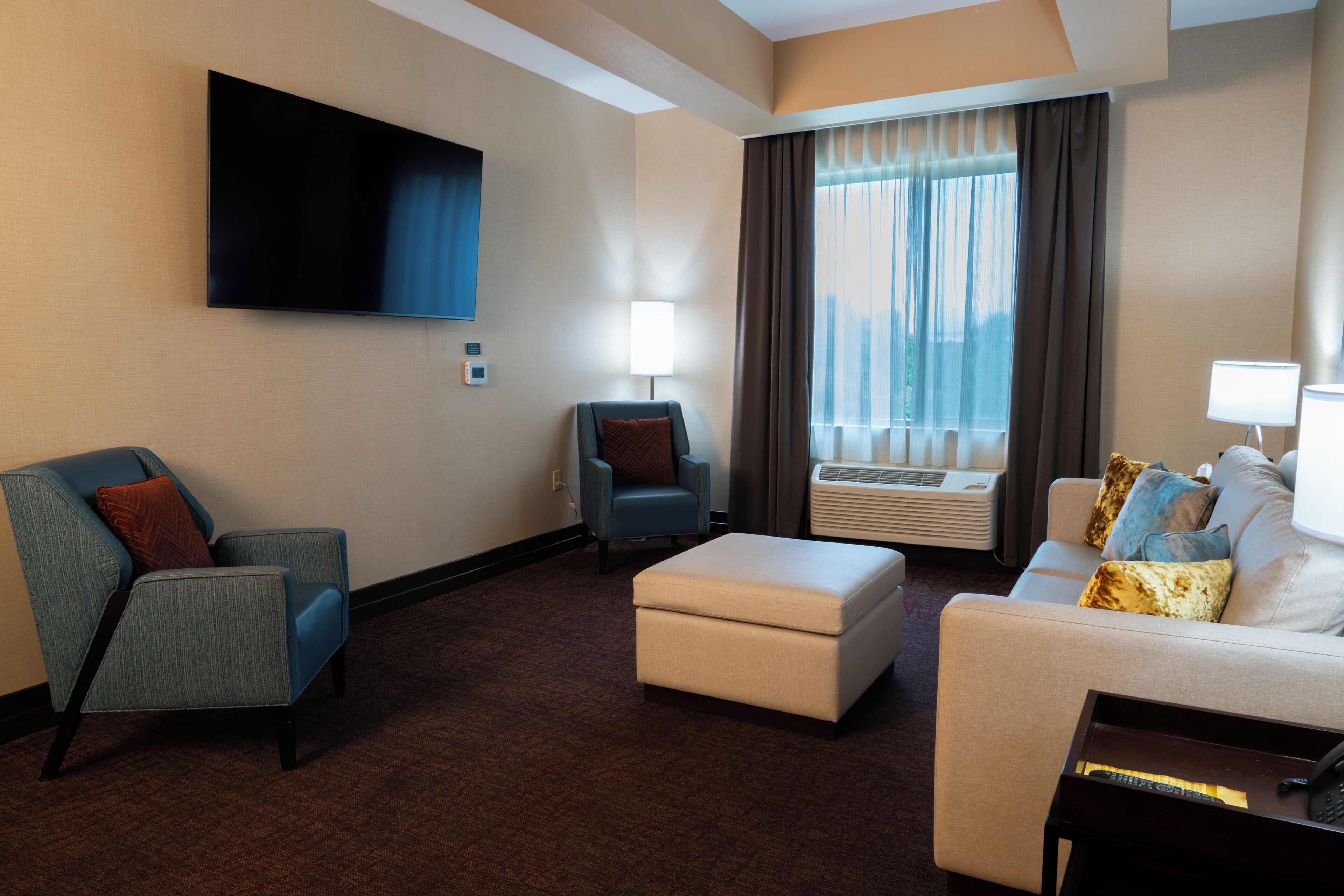A hotel room with a flat-screen TV, a sofa, a chair, an ottoman, and a window with curtains.