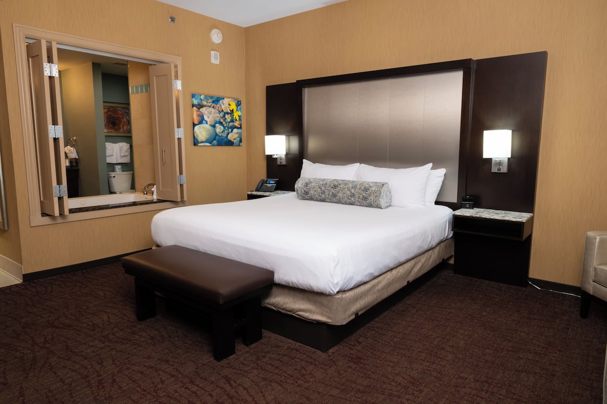 Habitación de hotel con una cama grande, un banco, una mesita de noche con lámpara, una mesita auxiliar y un espejo.