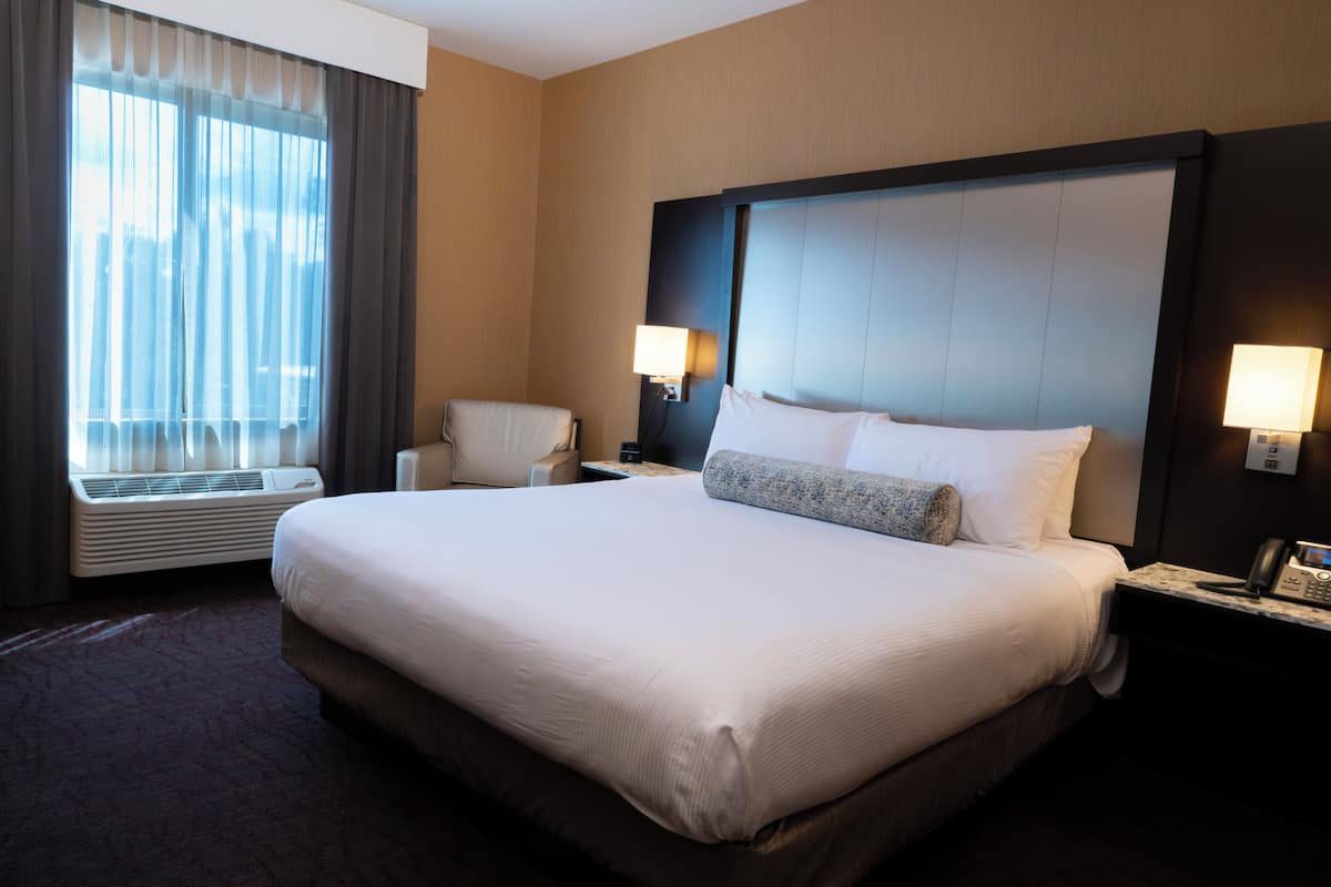 Habitación de hotel con una cama grande, dos mesitas de noche con lámparas, una silla, una ventana con cortinas y un teléfono instalado en la pared.