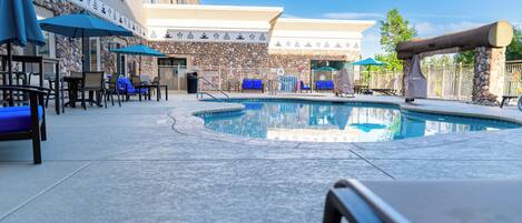 Zona de piscina con sillones de descanso y sombrillas, un muro de piedra y un edificio con un letrero.