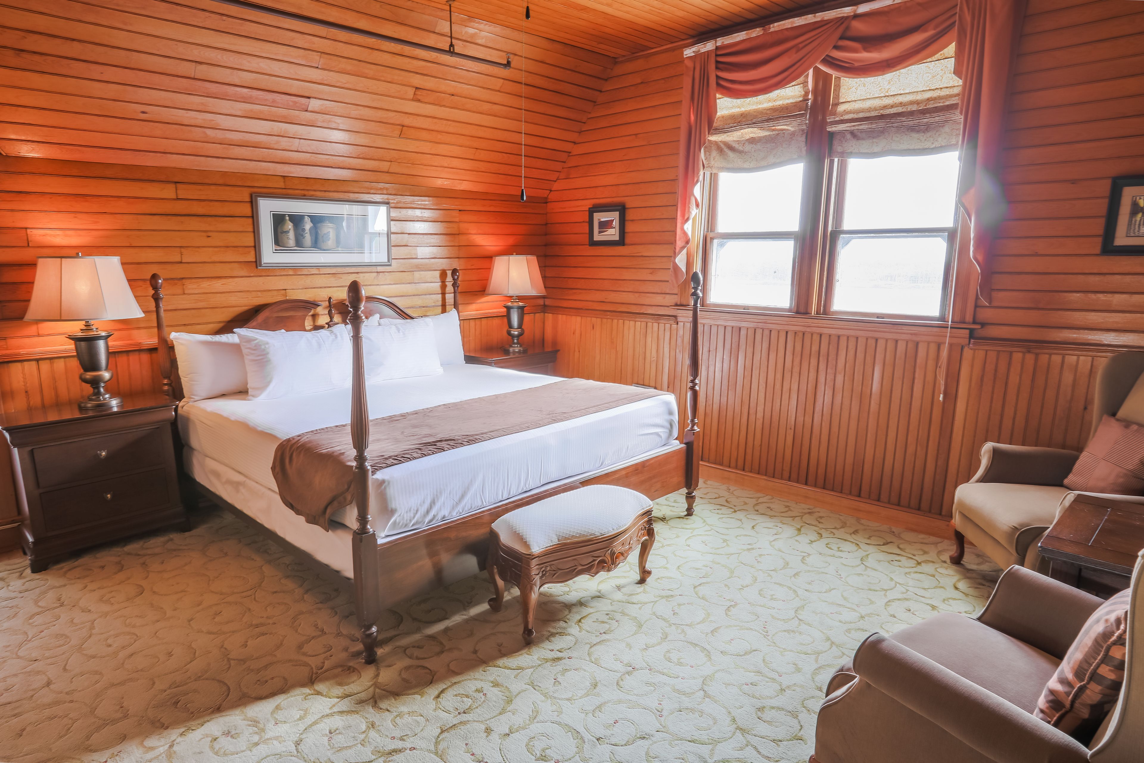 A wooden-paneled bedroom with a four-poster bed, two bedside lamps, a wooden bench, and a chair.
