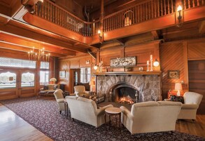 A spacious living room with a stone fireplace, wooden flooring, and a high ceiling. There are several upholstered chairs and a small round table.