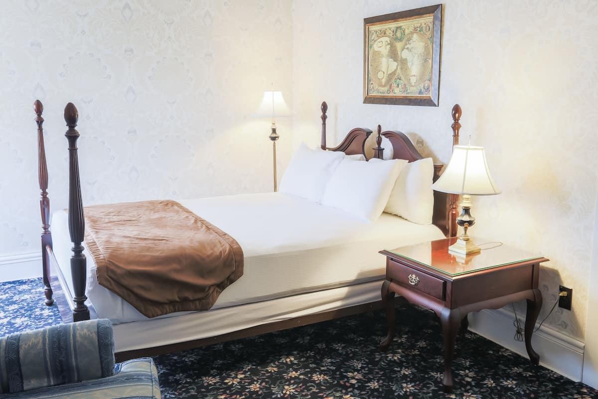 Un lit bien fait, recouvert d’un plaid marron, une table de chevet en bois avec une lampe, et un tableau encadré accroché au mur.