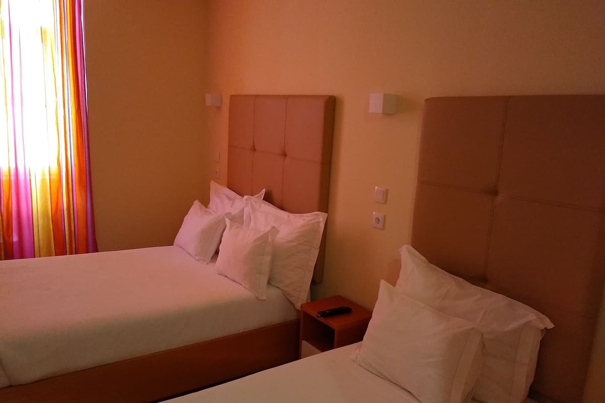 A hotel room with two beds, each with white bedding and pillows. There is a wooden nightstand between the beds with a remote control on it. The room has a beige wall and a window with curtains.