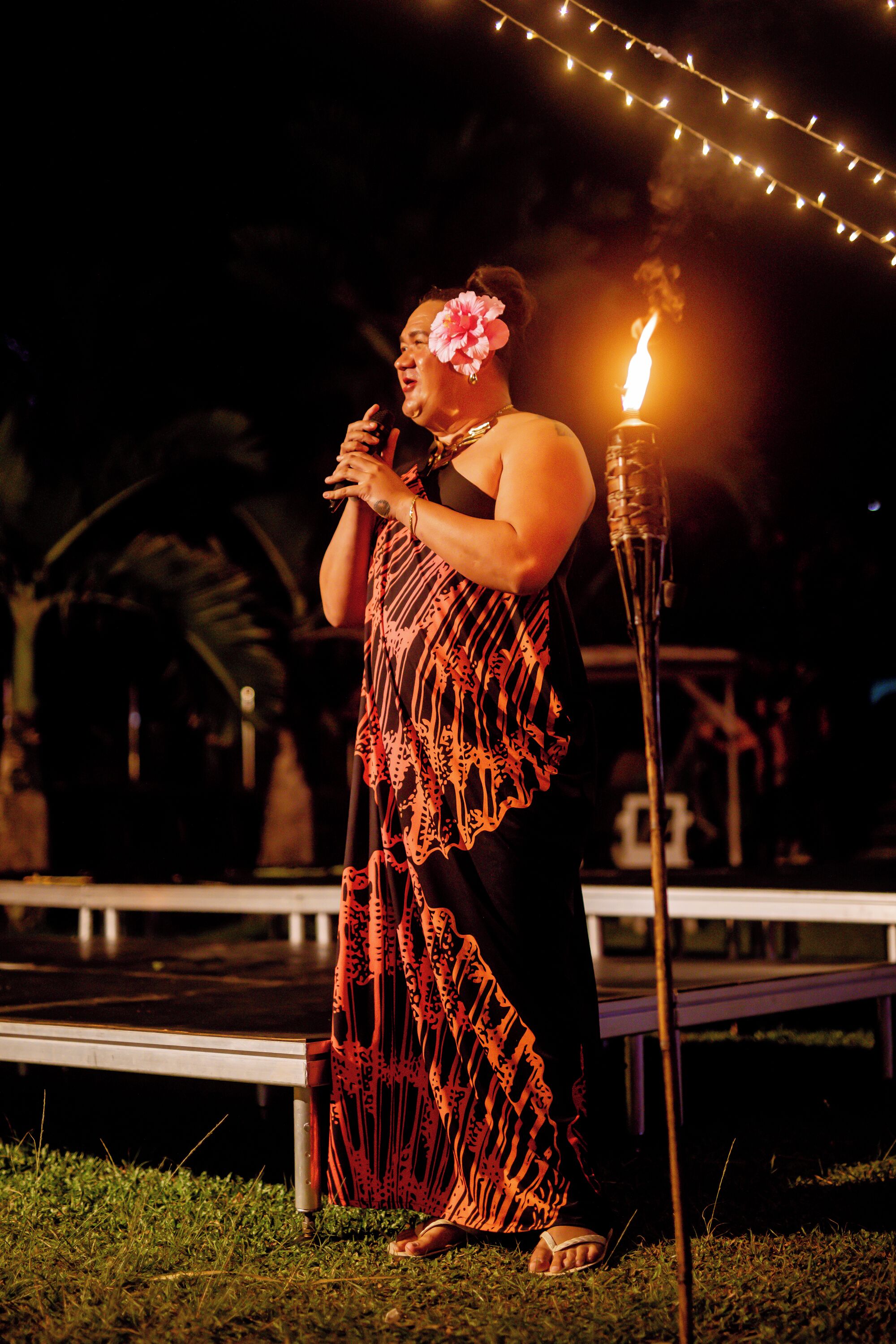 A person standing on a stage with a microphone, wearing a patterned dress, and a candle in the background.