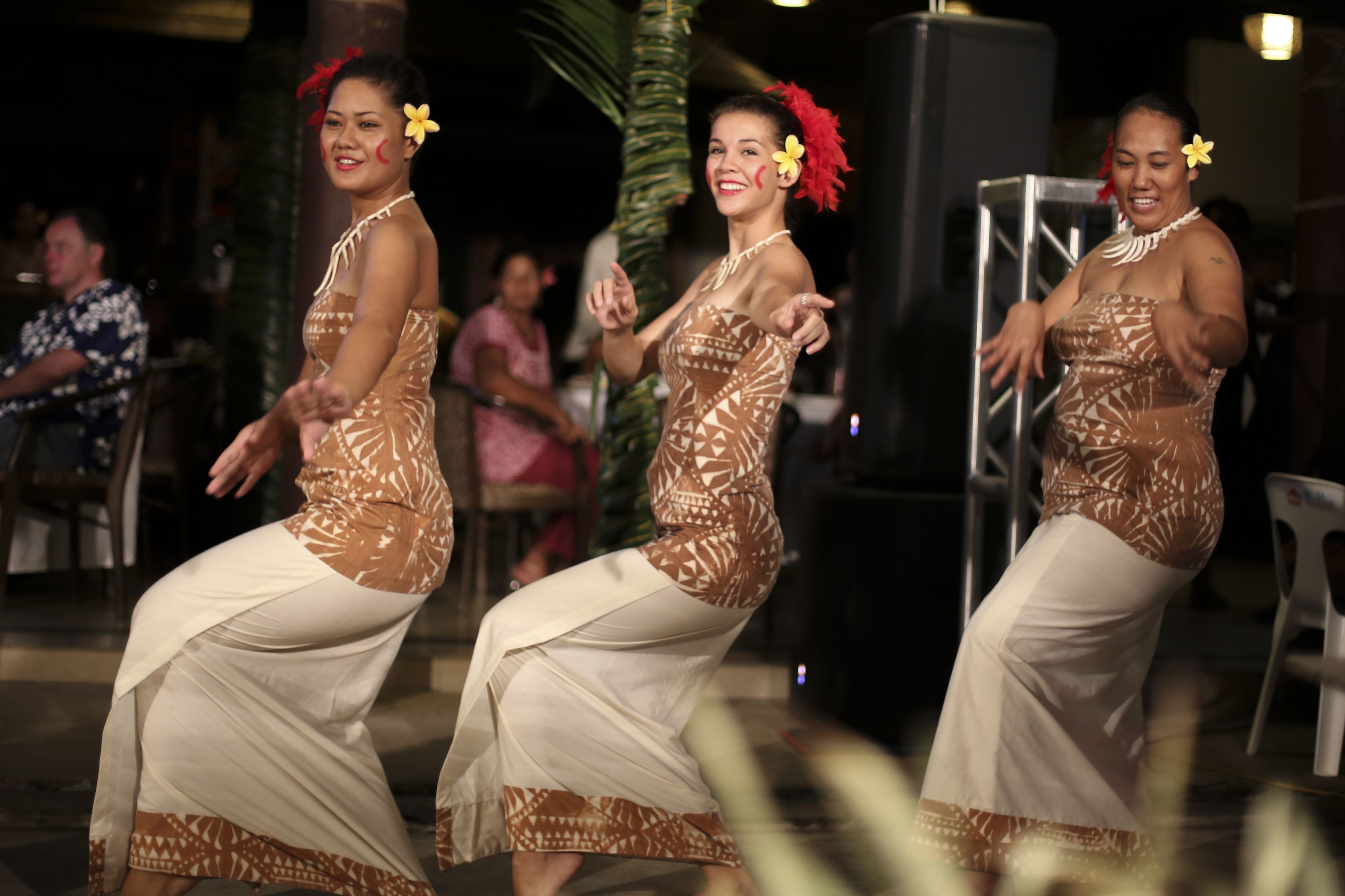 A group of people performing a dance in a casual outdoor setting with chairs and a palm tree in the background.