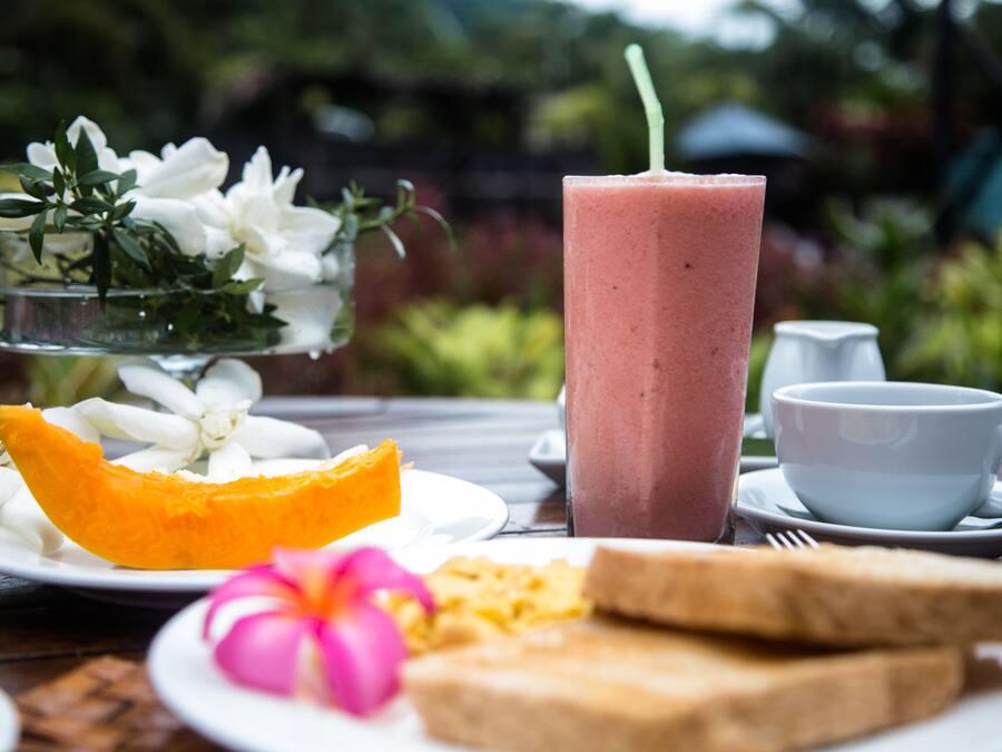 A table with a glass of pink smoothie, a plate with a slice of mango, a flower, and two slices of toast.