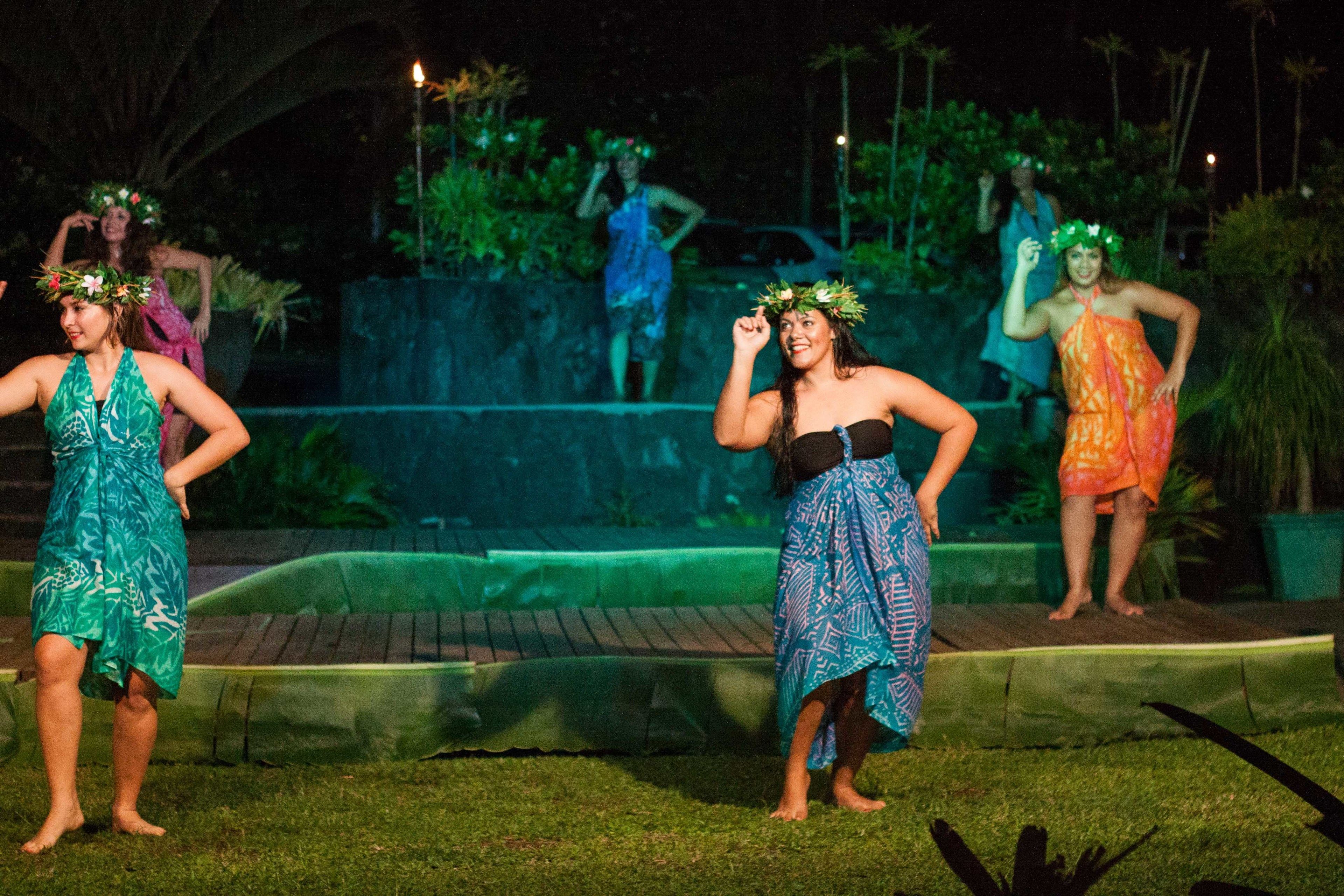 Un grupo de personas vestidas con ropa tropical se presenta en un escenario al aire libre, con un muro de ladrillo y follaje en el fondo.
