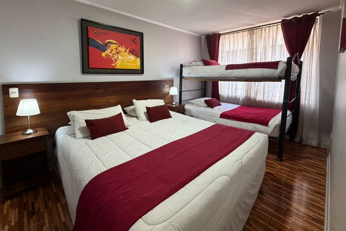 Habitación de hotel con dos camas, una litera, cabecero de madera, mesita de noche con lámpara, un cuadro en la pared y una ventana con cortinas.