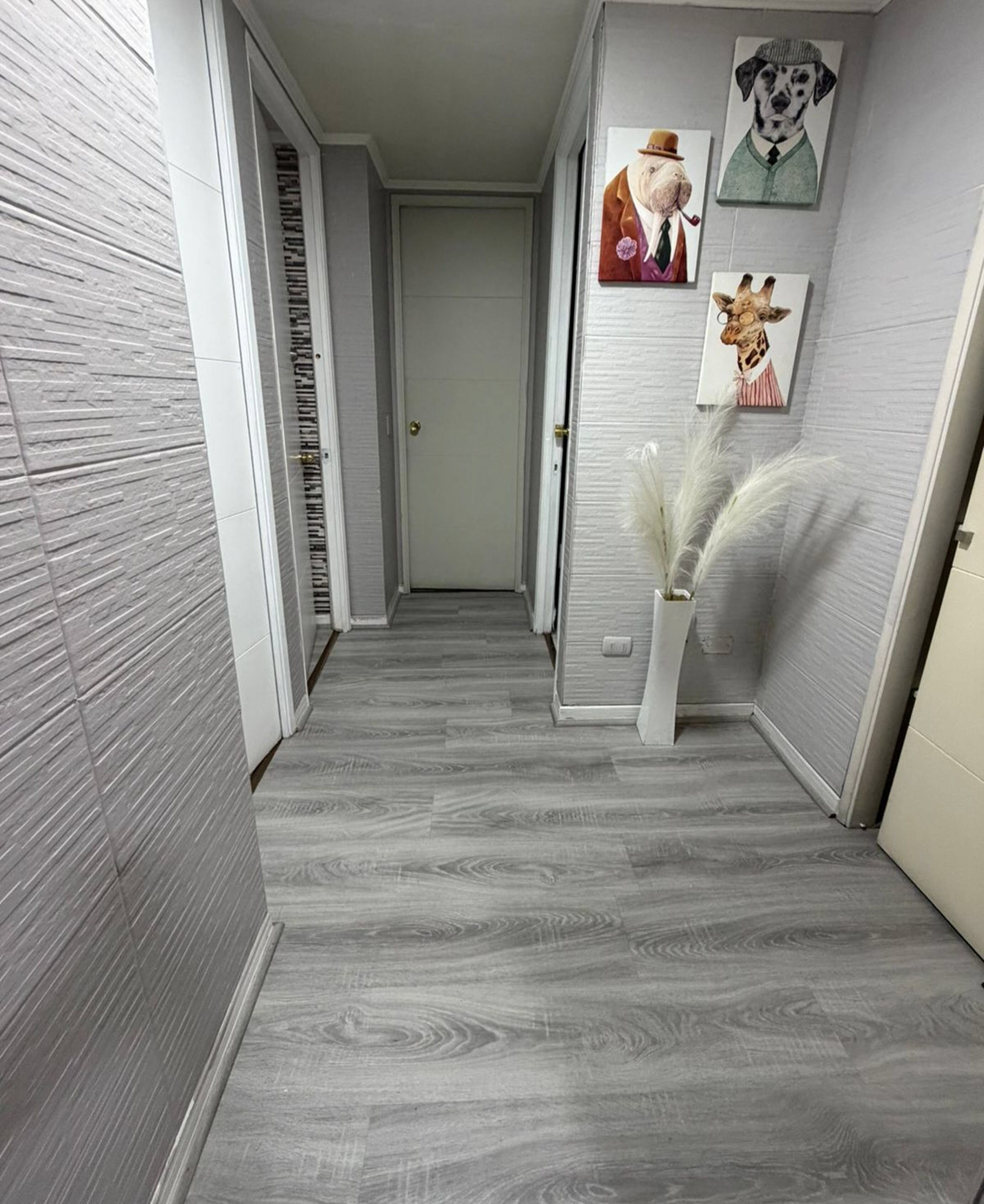 Un couloir avec un plancher en bois gris, des murs blancs et une porte au fond. Des tableaux encadrés ornent le mur.