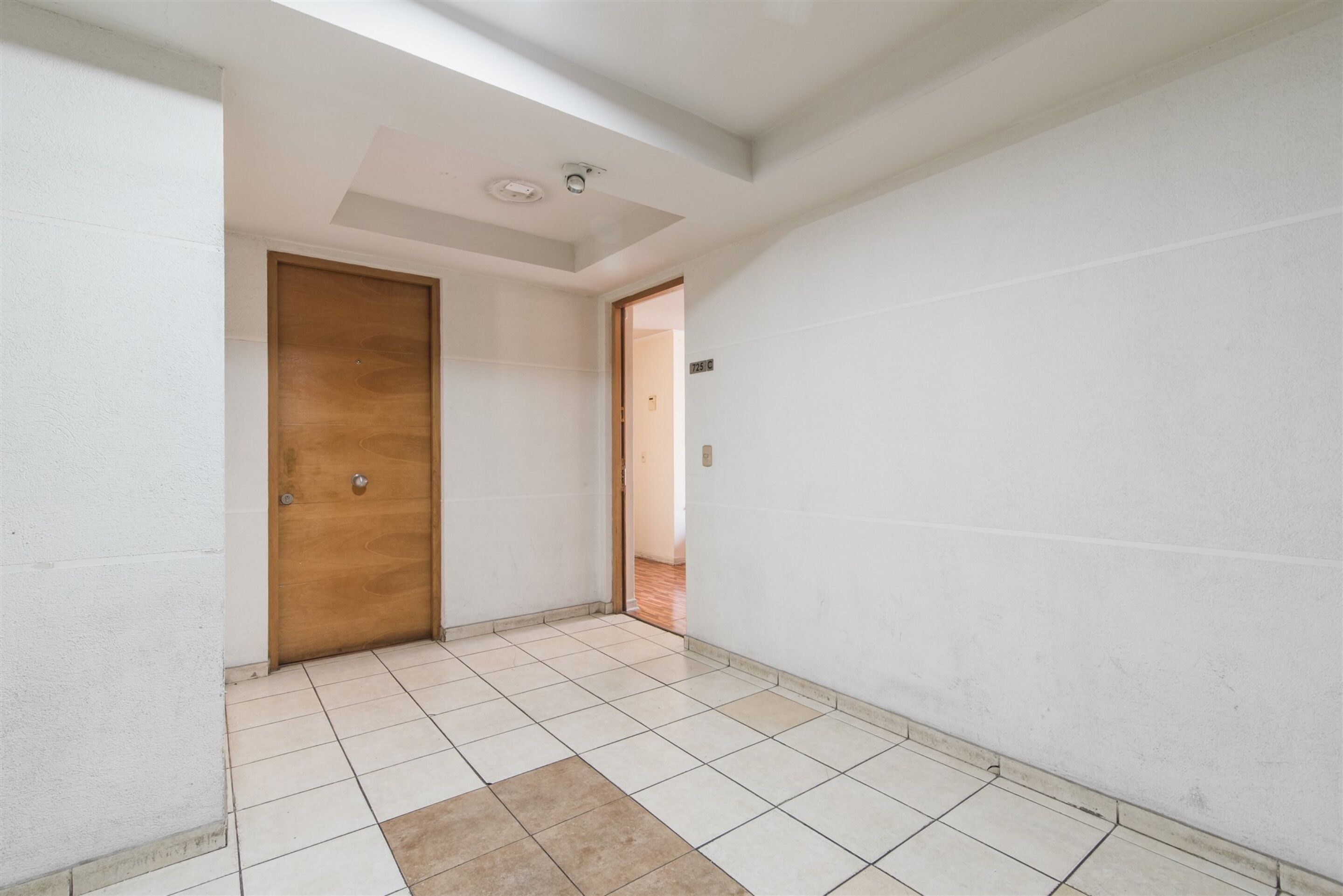 Un pasillo con puerta de madera, paredes blancas y suelo de baldosas.