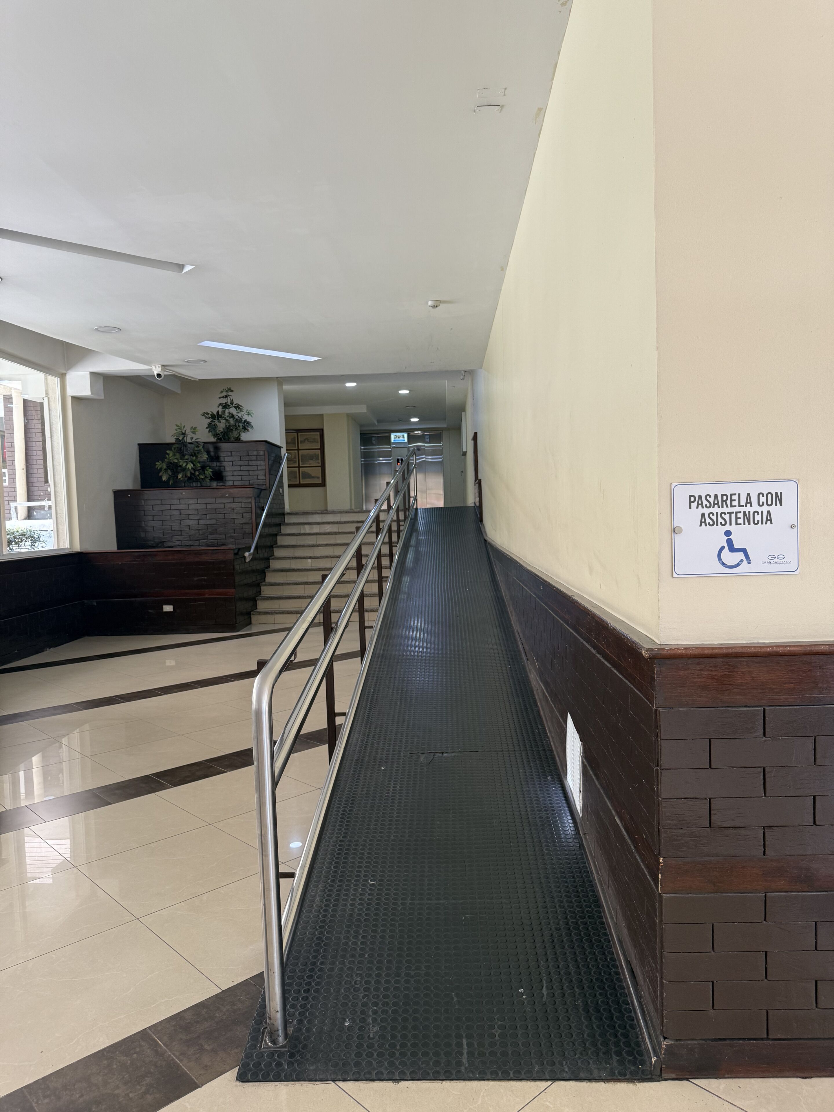 Un couloir avec une rampe menant à un ascenseur, un panneau indiquant la présence d’un service d’assistance pour les personnes en fauteuil roulant, et un escalier muni d’une main courante.