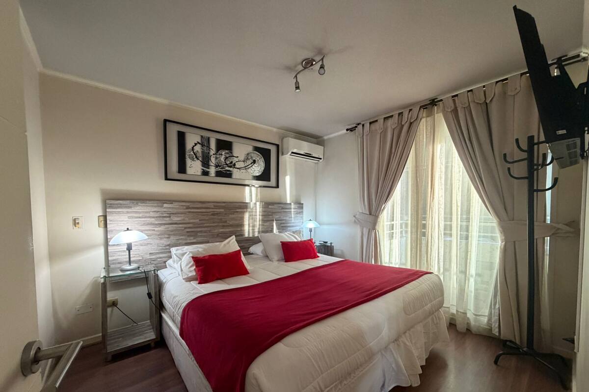 Una habitación de hotel con cama, mesita de noche, lámpara, ventilador de techo, televisor y ventana con cortinas.