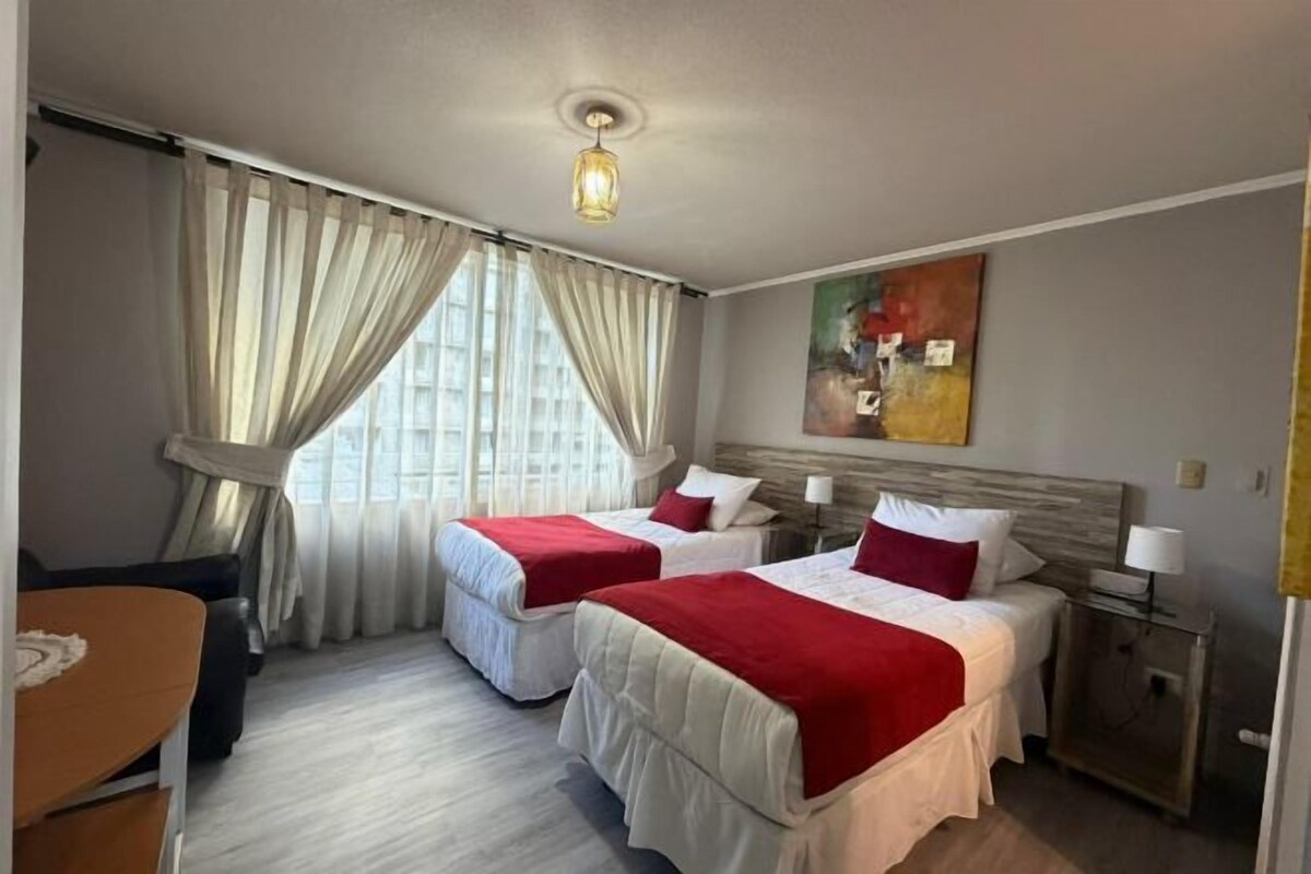 A hotel room with two beds, a desk, a chair, a nightstand, and a painting on the wall.