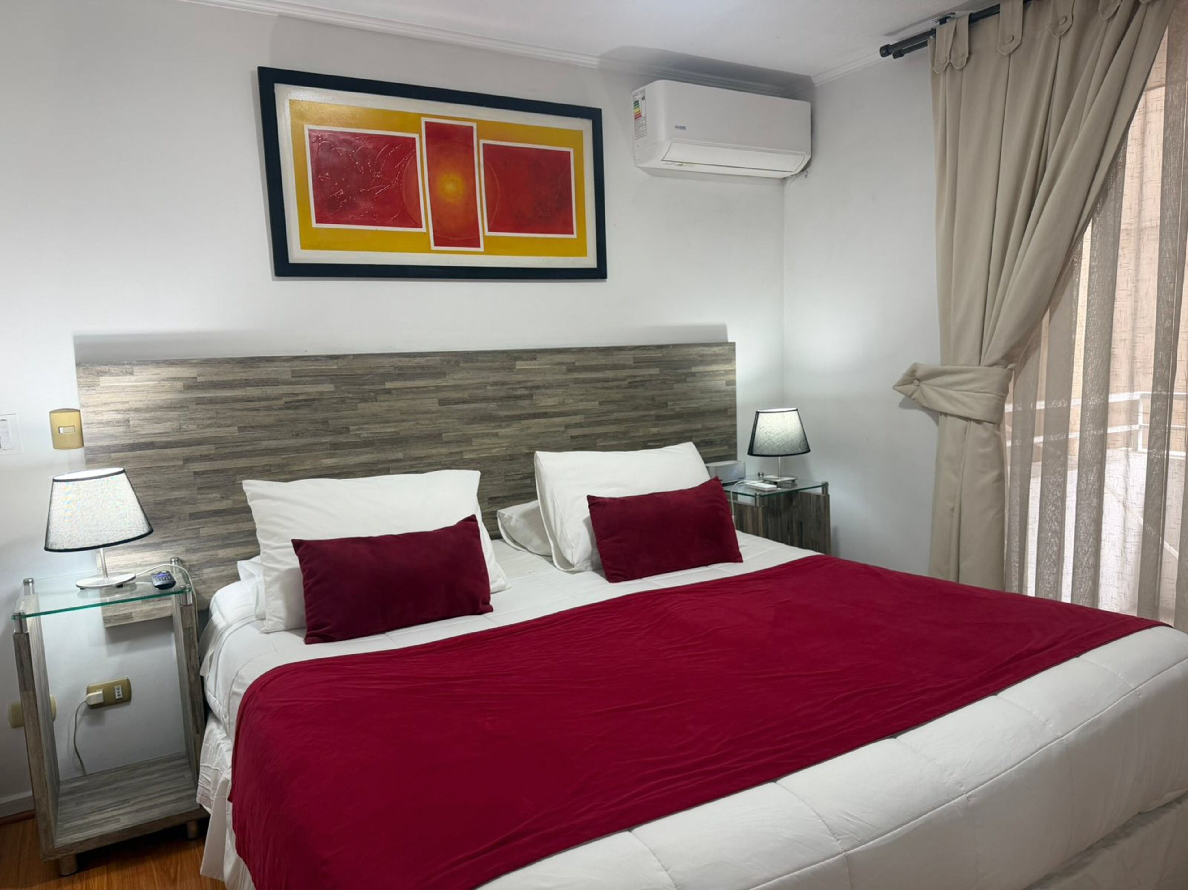 Une chambre d’hôtel comprenant un lit, deux tables de chevet, un climatiseur fixé au mur et une fenêtre avec des rideaux.