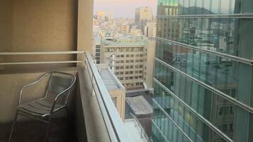 A balcony with a metal chair, a glass building to the right, and a cityscape in the background.