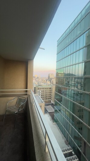 A balcony with a metal chair, a glass building to the right, and a cityscape in the background. - VR Suite (Santiago)