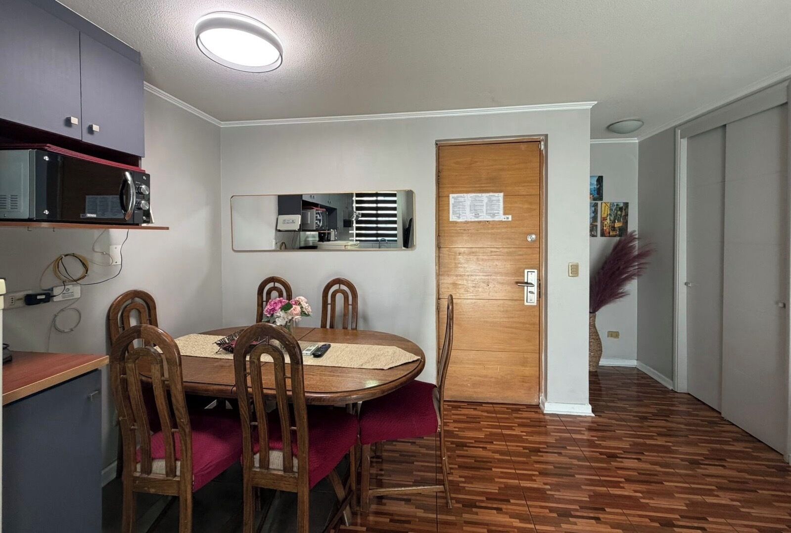 A dining area with a wooden table, four chairs, a microwave, a refrigerator, and a door with a bulletin board.