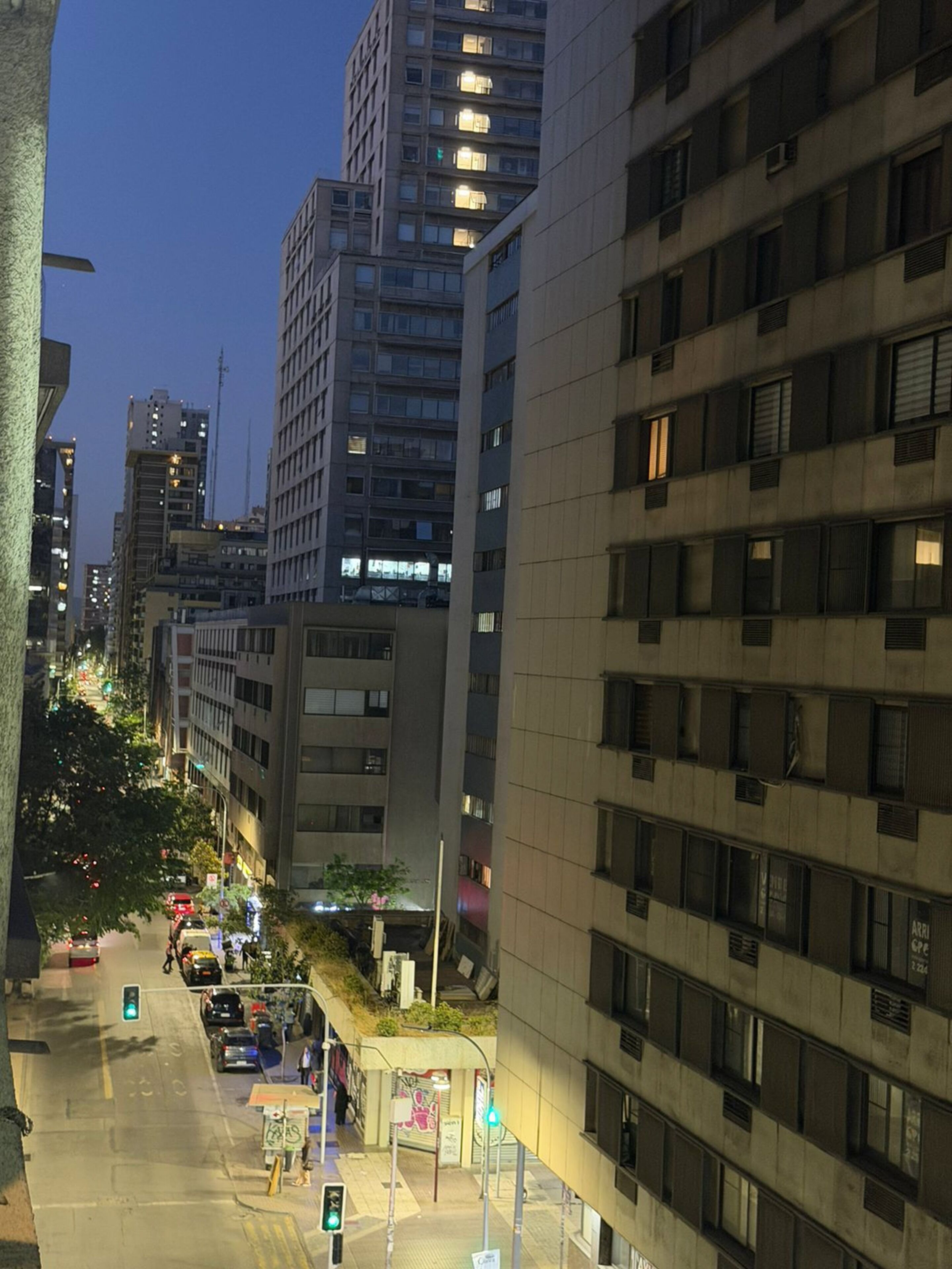 A city street at dusk with modern buildings, a pedestrian crossing, and street lamps.