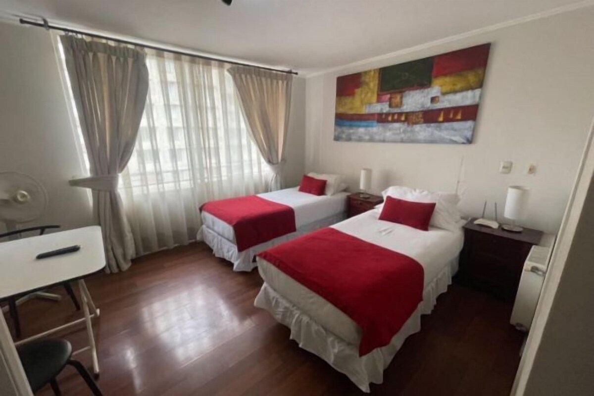Habitación de hotel con dos camas, un escritorio, una silla, una mesita de noche y un cuadro en la pared.