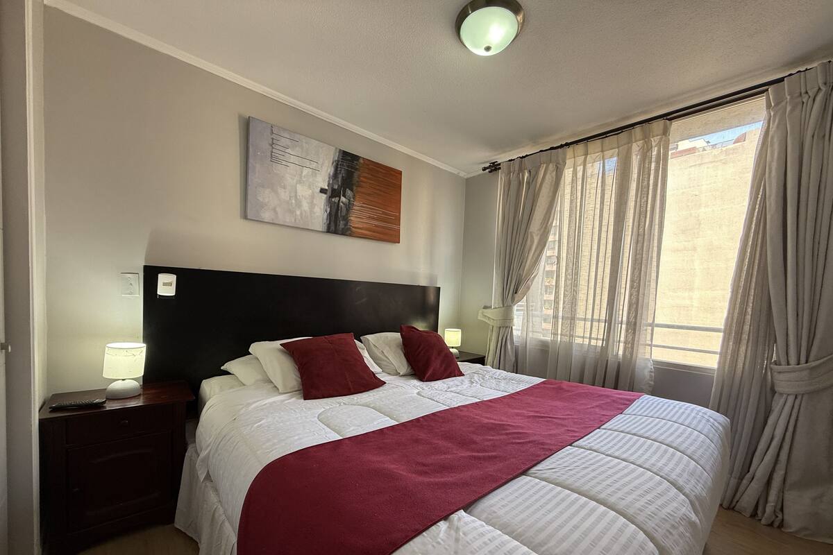 Una habitación de hotel con una cama grande, una mesita de noche con una lámpara, un cuadro en la pared y un ventanal con cortinas.
