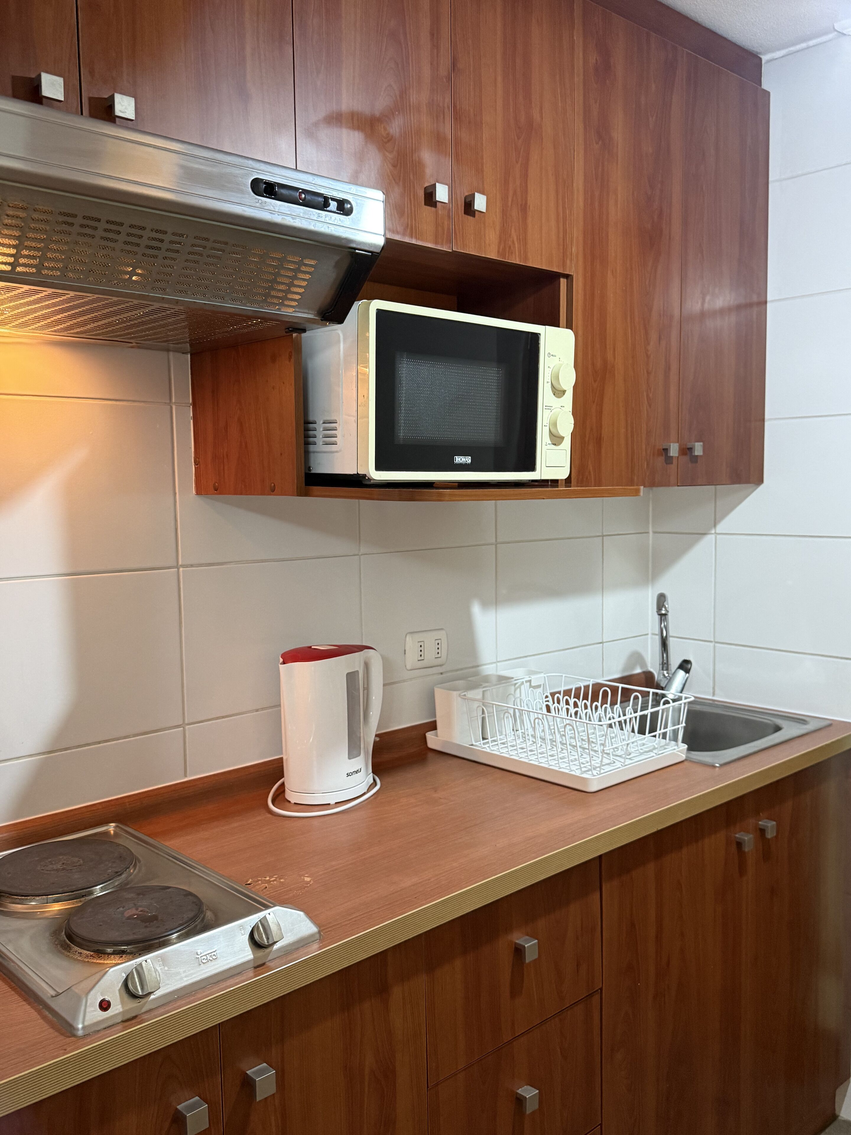 A kitchen with wooden cabinets, a microwave, a kettle, a sink, and a stove.