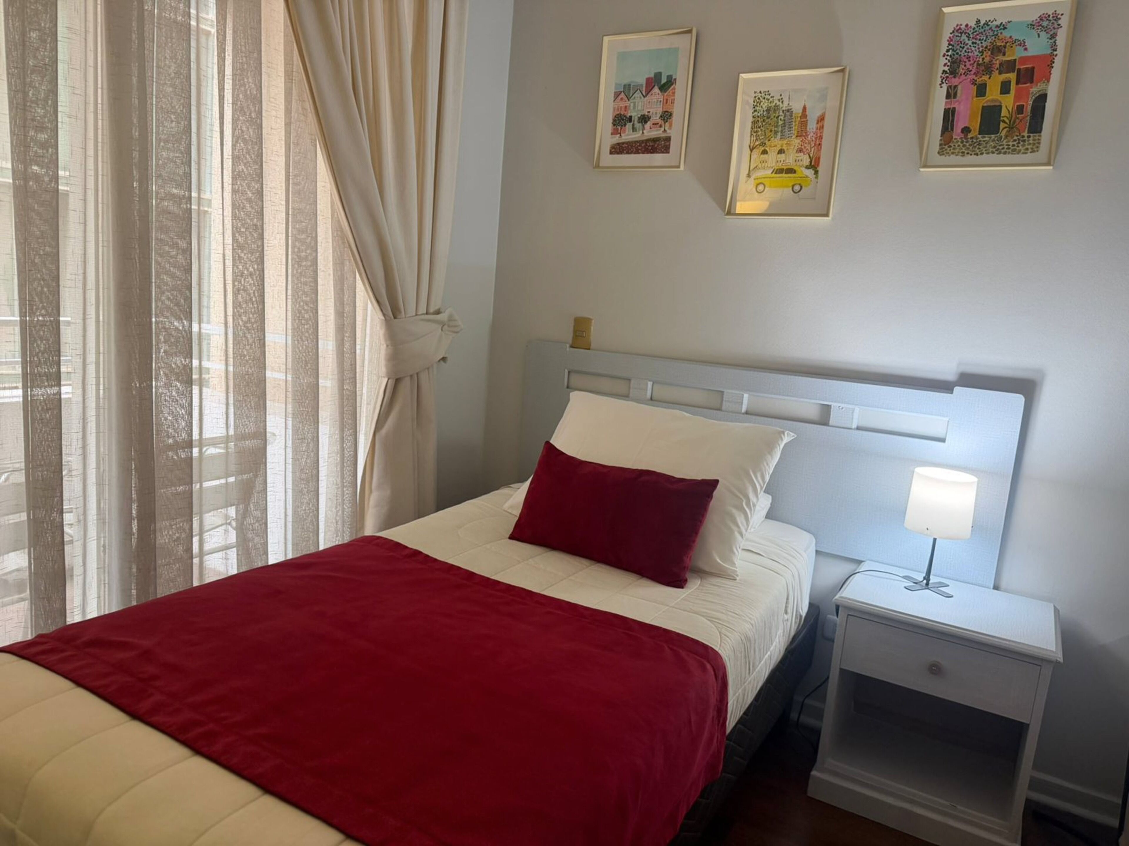 A neatly made bed with a red blanket, a white pillow, and a nightstand with a lamp. There are three framed artworks on the wall.