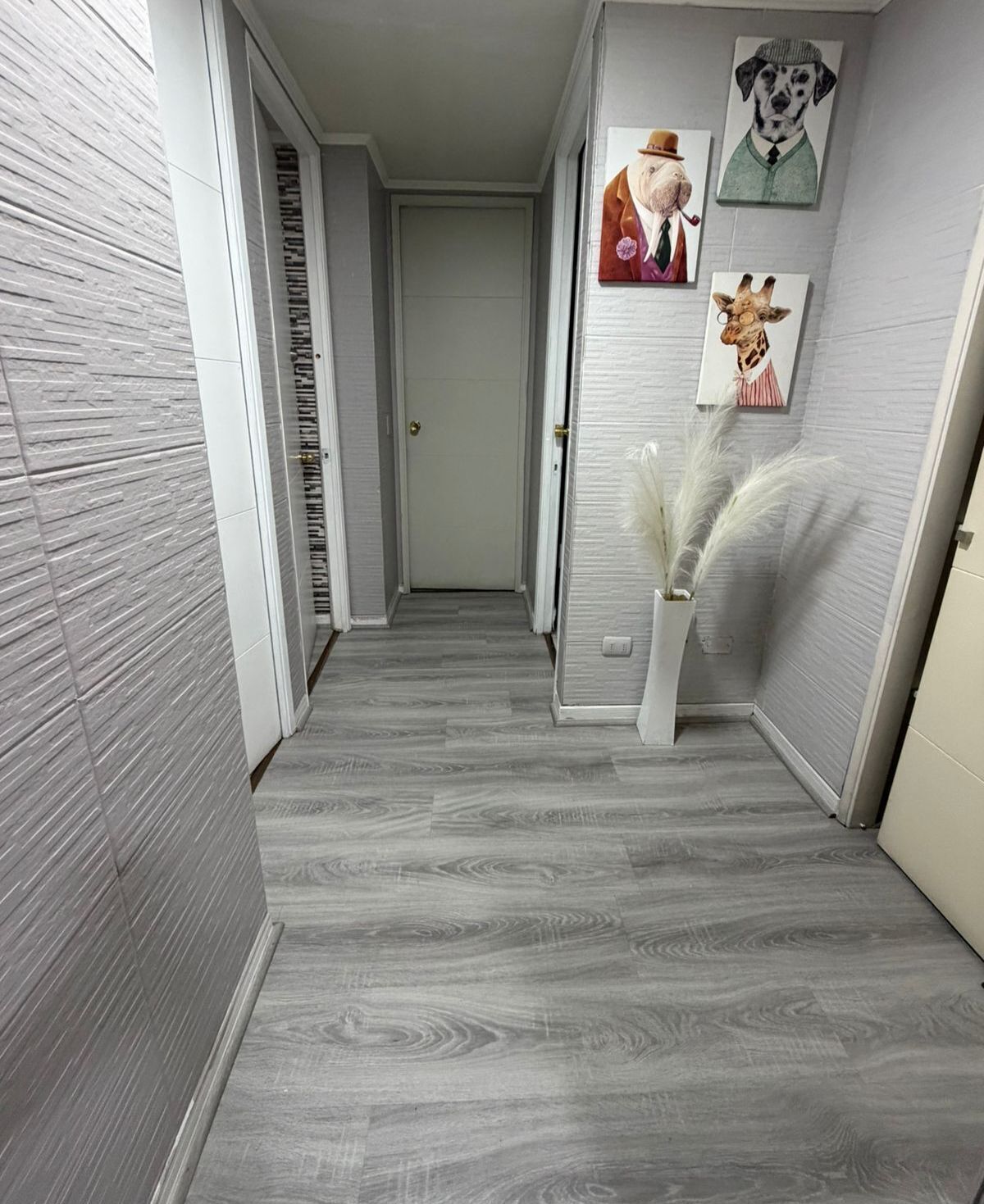 Un pasillo con piso de madera gris, paredes blancas y una puerta al final. Hay cuadros enmarcados en la pared y un florero con flores blancas.
