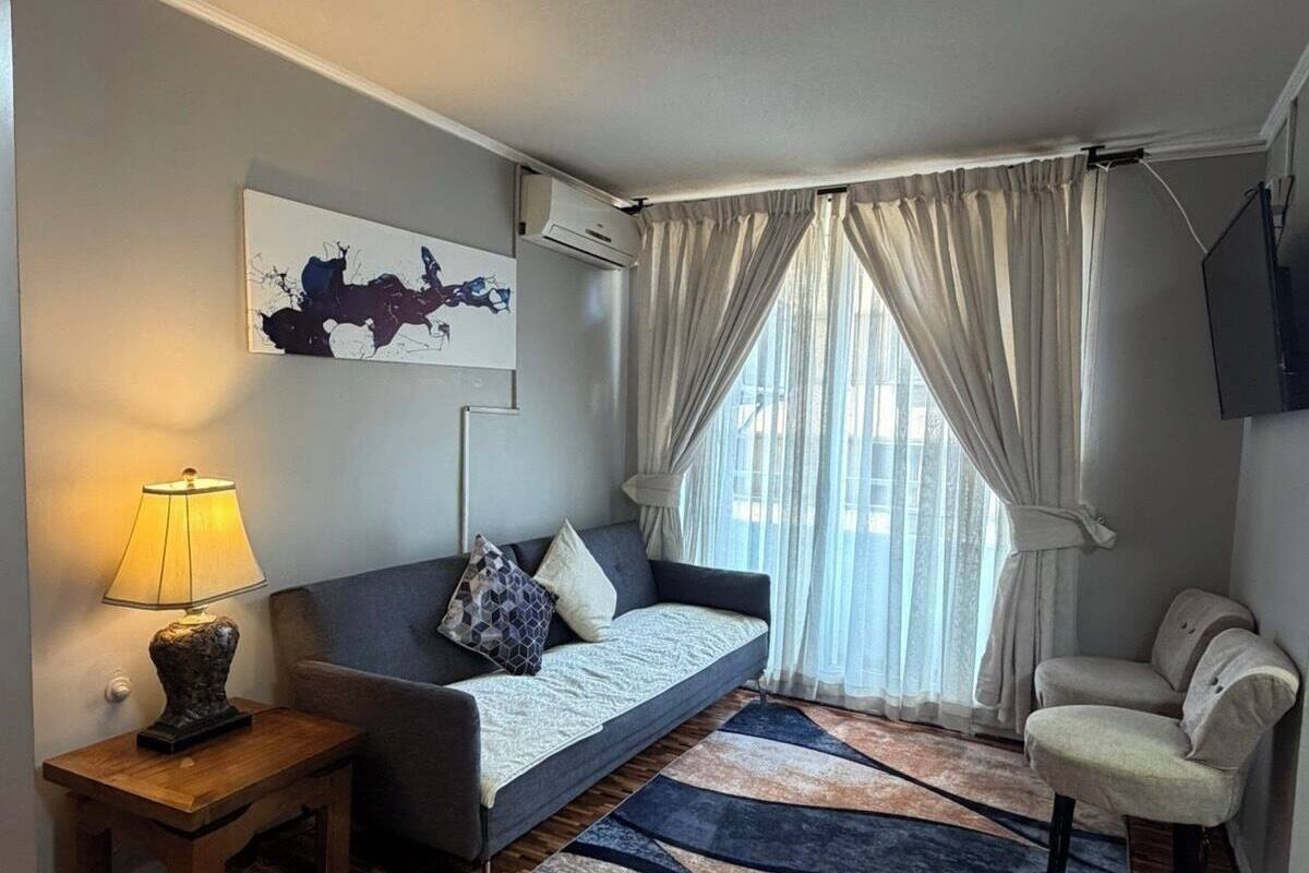 Una sala de estar moderna con un sofá, una mesita auxiliar con una lámpara, un tapete estampado y un ventanal con cortinas.