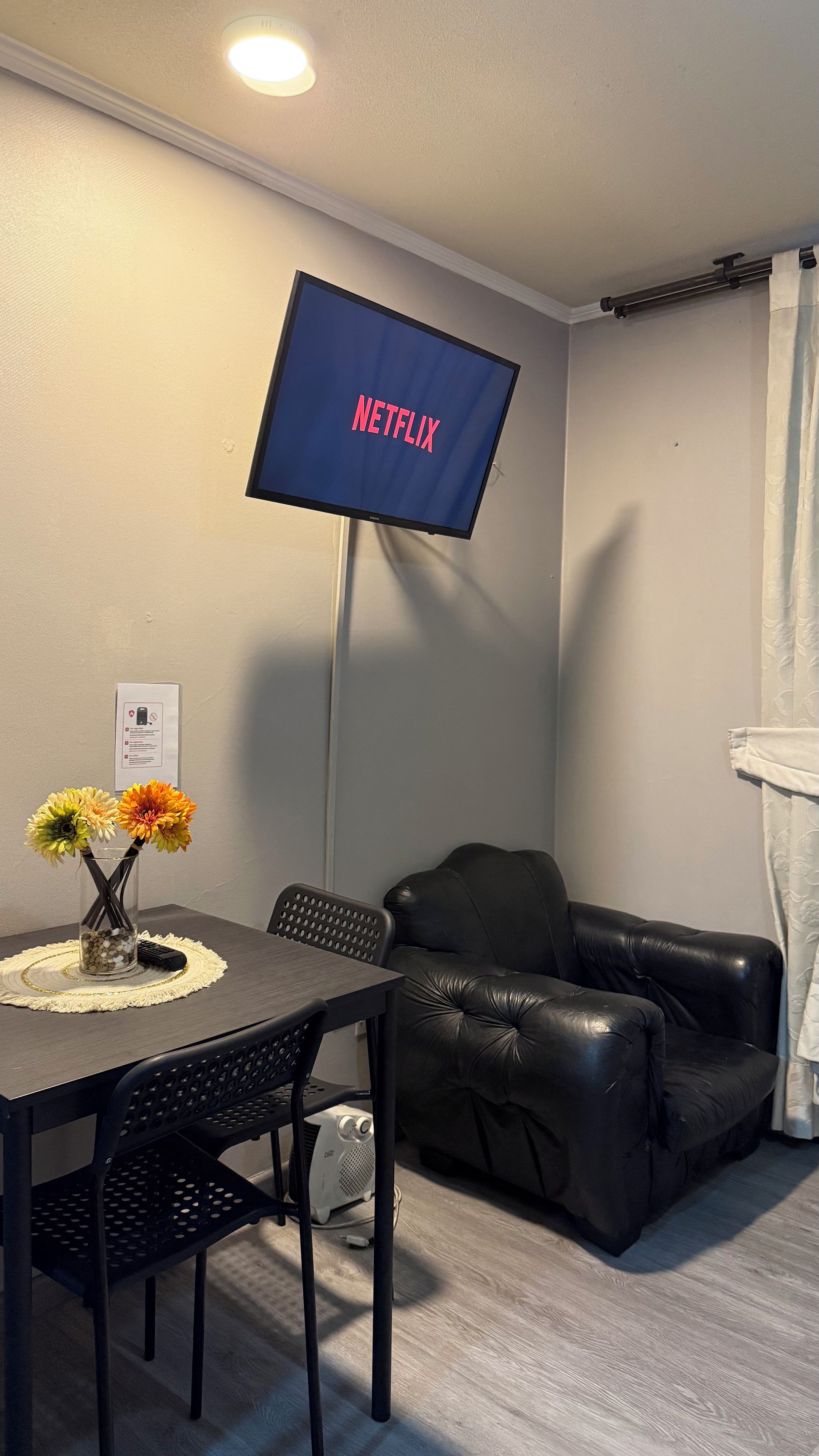 A room with a flat-screen TV displaying the Netflix logo, a dining table with chairs, a black leather sofa, and a small decorative vase with flowers.