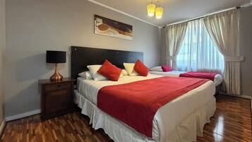 Quarto de hotel com duas camas, uma mesinha de cabeceira, uma luminária, um quadro na parede e uma janela com cortinas.