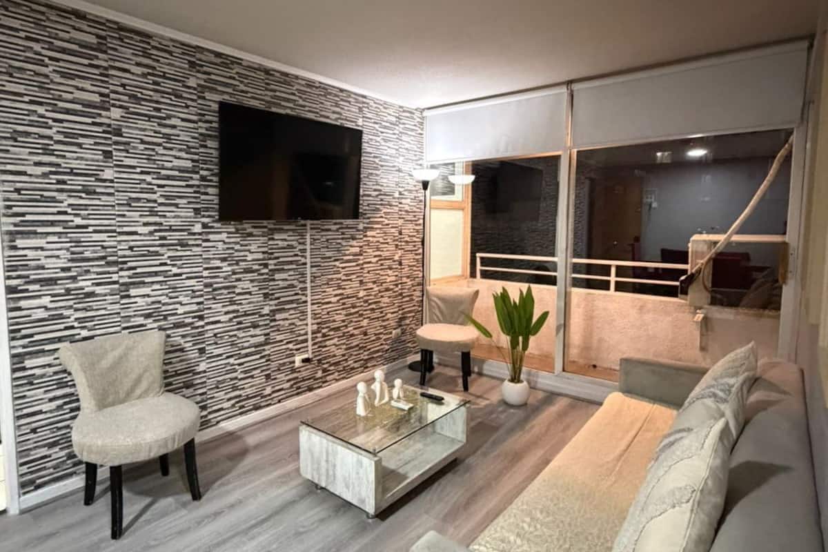 Una sala moderna con pared de diseño de piedra, televisor de pantalla plana, mesa de centro de vidrio, sofá color beige y una planta en maceta.