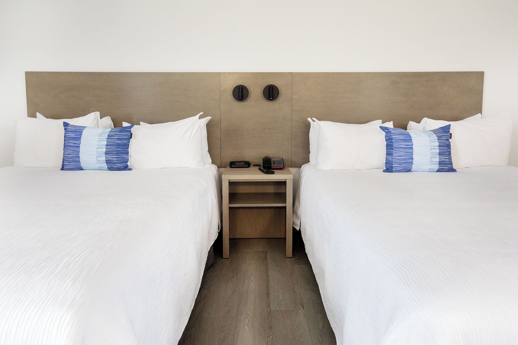 A hotel room with two beds, each with white bedding and blue striped pillows. There is a wooden nightstand between the beds with a telephone and a small table.