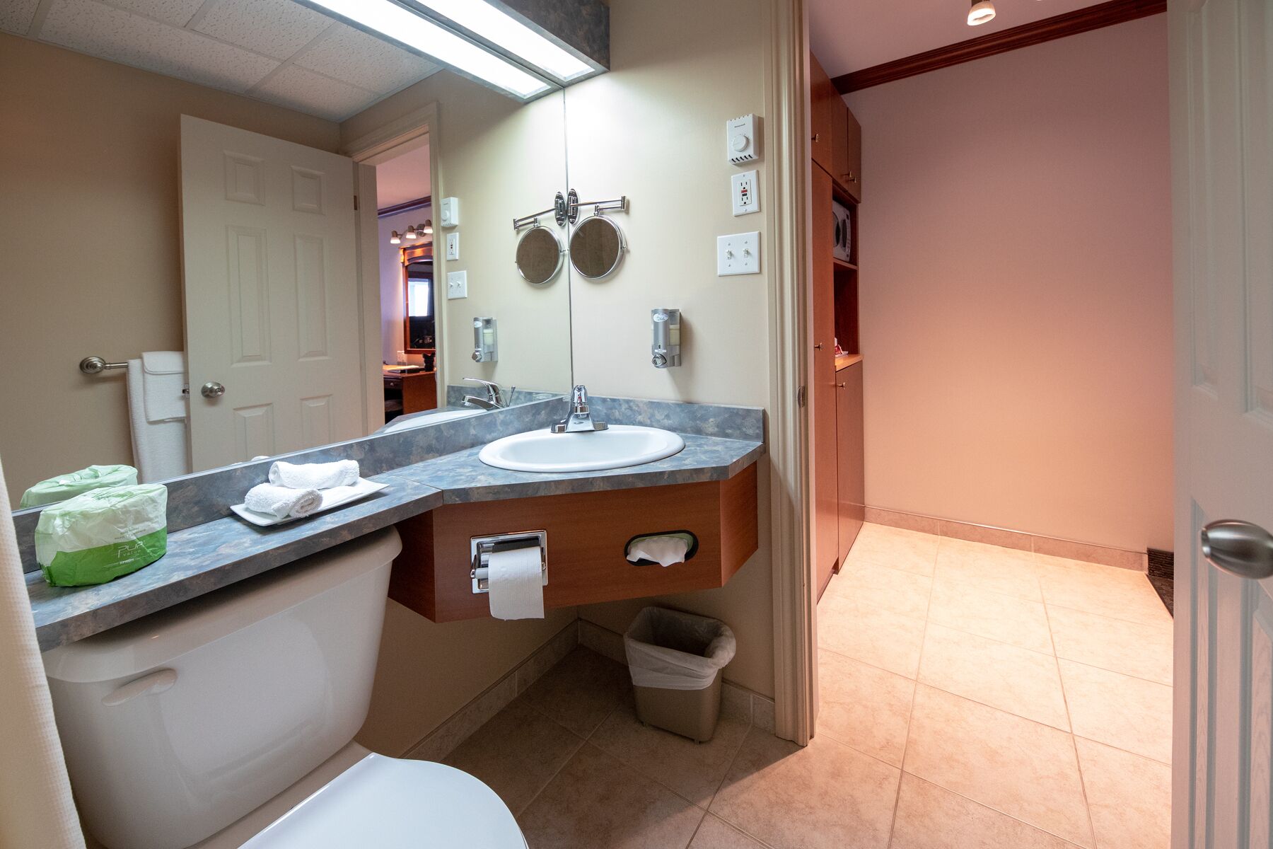 A bathroom with a sink, toilet, and a mirror. There are towels and a trash can visible.