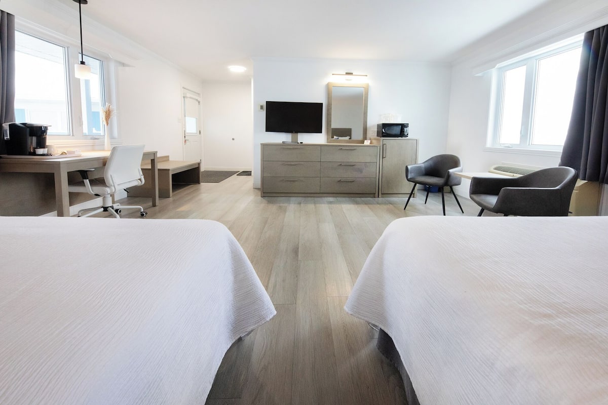 A hotel room with two beds, a desk, a chair, a dresser, a television, and a large window.