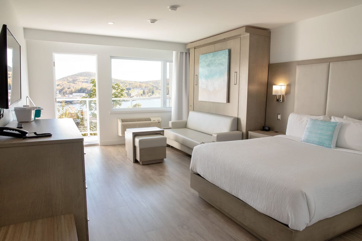 A hotel room with a large bed, a desk with a computer, a chair, a sofa, a window with a view of a lake, and a painting on the wall.