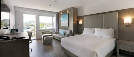 Habitación de hotel moderna con una cama grande, un escritorio y un sofá. Se ve el exterior a través de las ventanas.