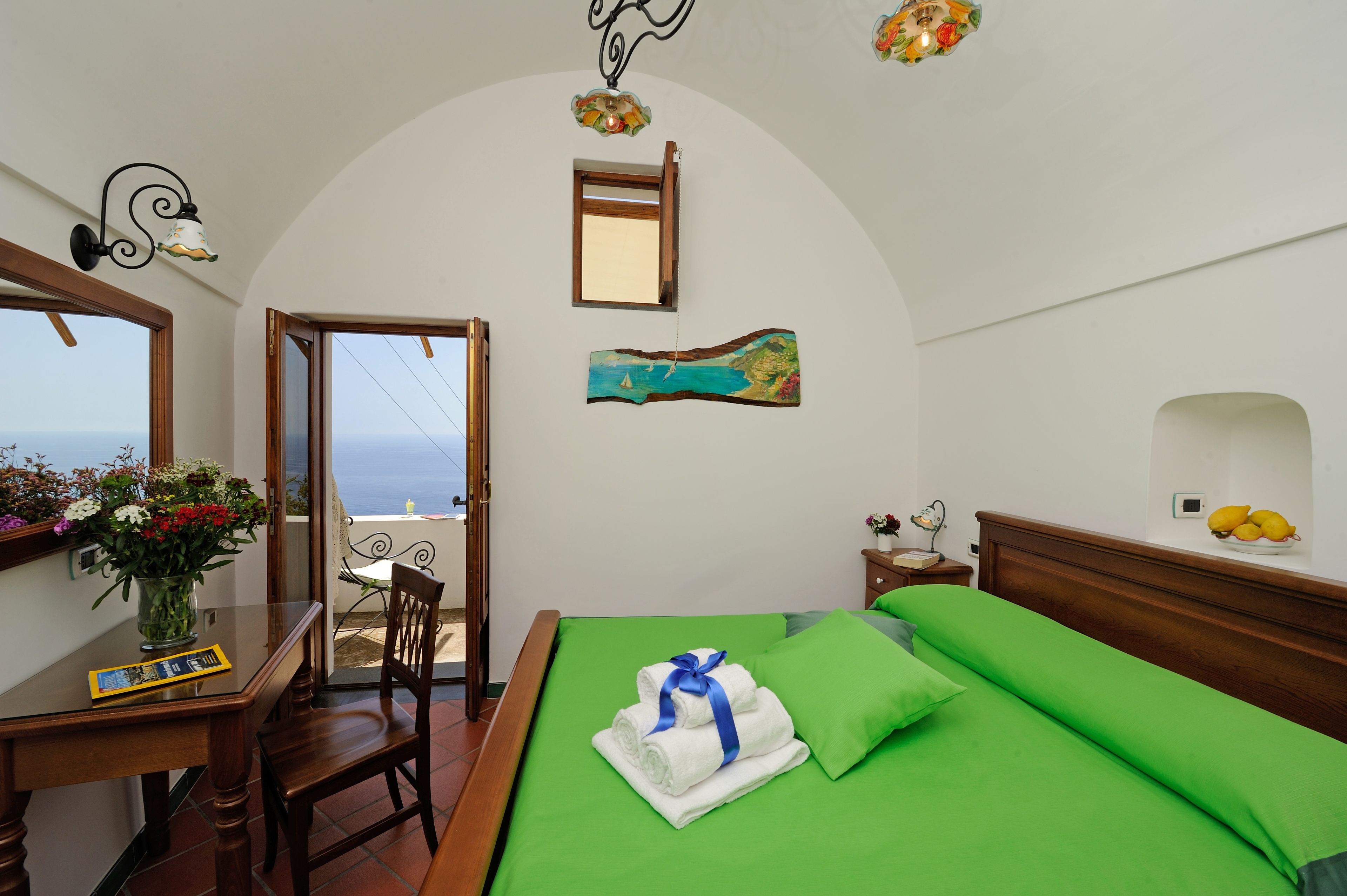 A bedroom with a bed, a desk with a vase of flowers, a chair, a window with a view, and a painting on the wall.