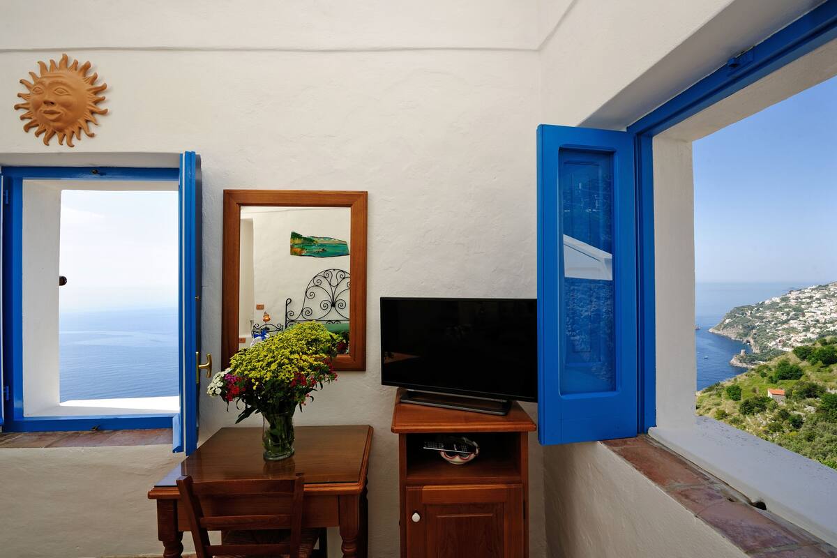 A room with a wooden table, a TV, and a vase of flowers. There is a view of the sea through an open door and a window.