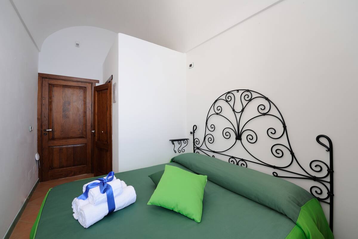A hotel room with a wooden door, a metal headboard with a decorative design, a green bedspread, and a green pillow.