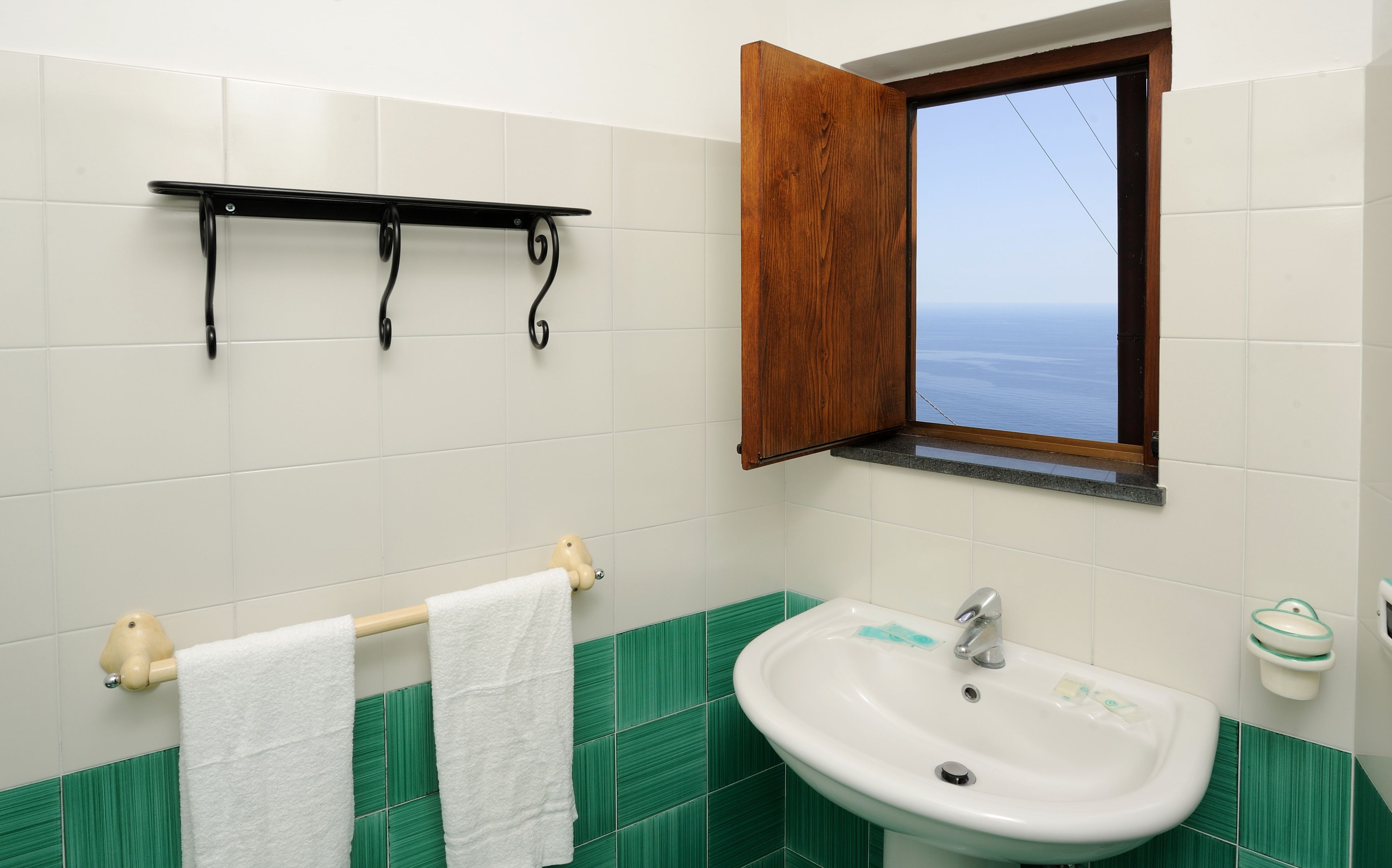 Une salle de bain avec un lavabo blanc, des carreaux verts, un porte-serviettes avec des serviettes blanches, un meuble à pharmacie en bois et une fenêtre donnant sur la mer.