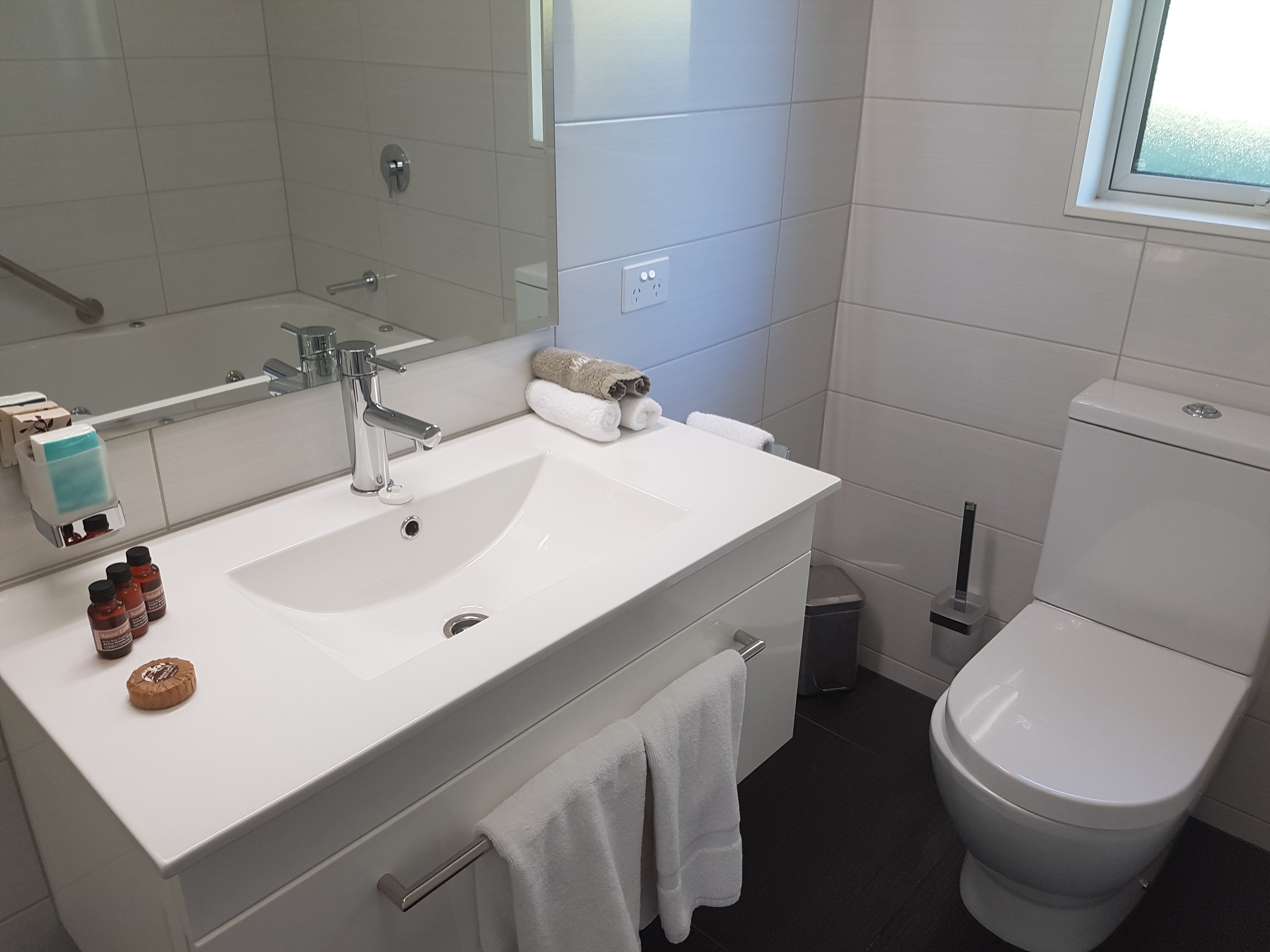 Casa de banho com lavatório branco, espelho e sanita. Há produtos de higiene pessoal no lavatório e um porta-toalhas com toalhas.
