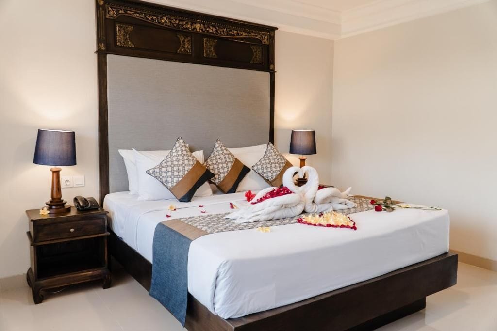 Habitación de hotel con una cama grande, cabecera de madera oscura, dos mesitas de noche con lámparas y almohadones decorativos.